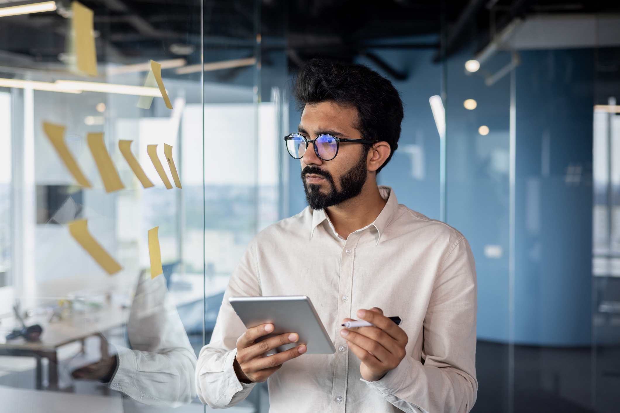 Direct Hire Dallas TX - Young Indian man working in office with tablet, standing near glass board with notes, processing data, thinking about project.