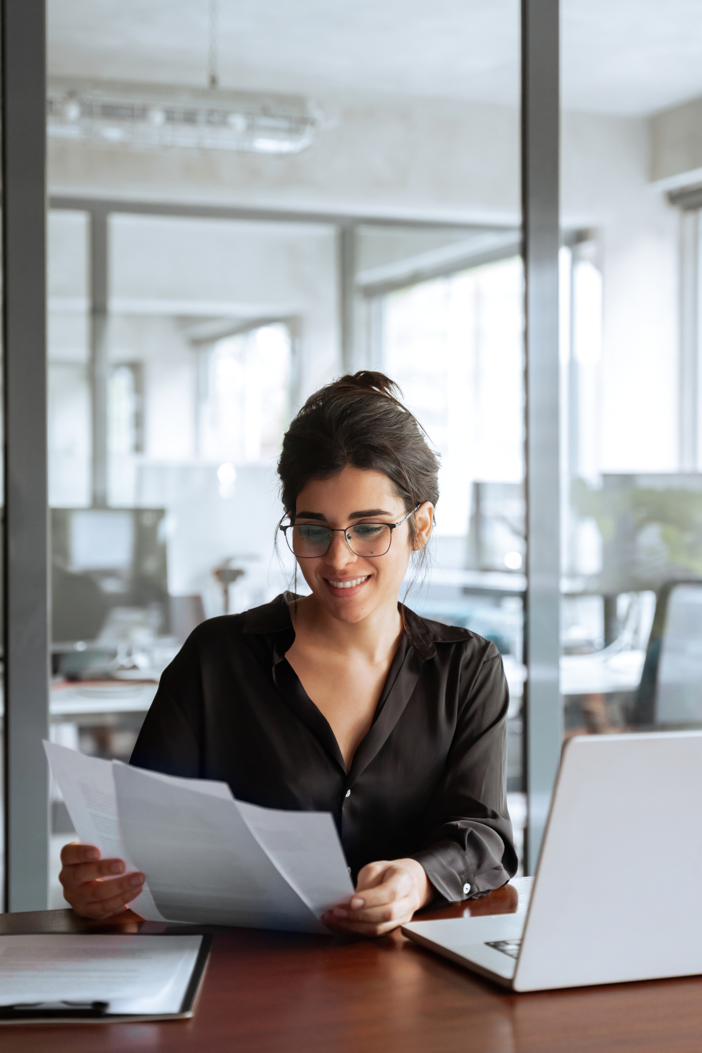 Executive Recruiters Dallas TX - Vertical smiling portrait business woman accountant holding documents, working at laptop pc computer doing online tax invoice, financial report. Latina businesswoman manager with paperwork in office